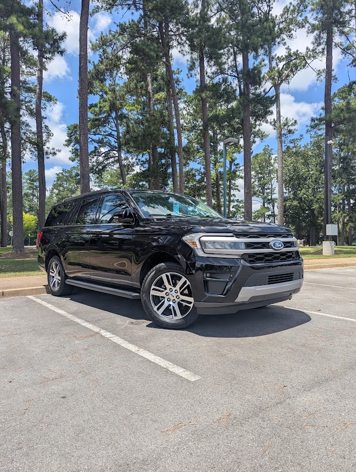 A black Ford Expedition SUV parked in a paved lot surrounded by tall pine trees.