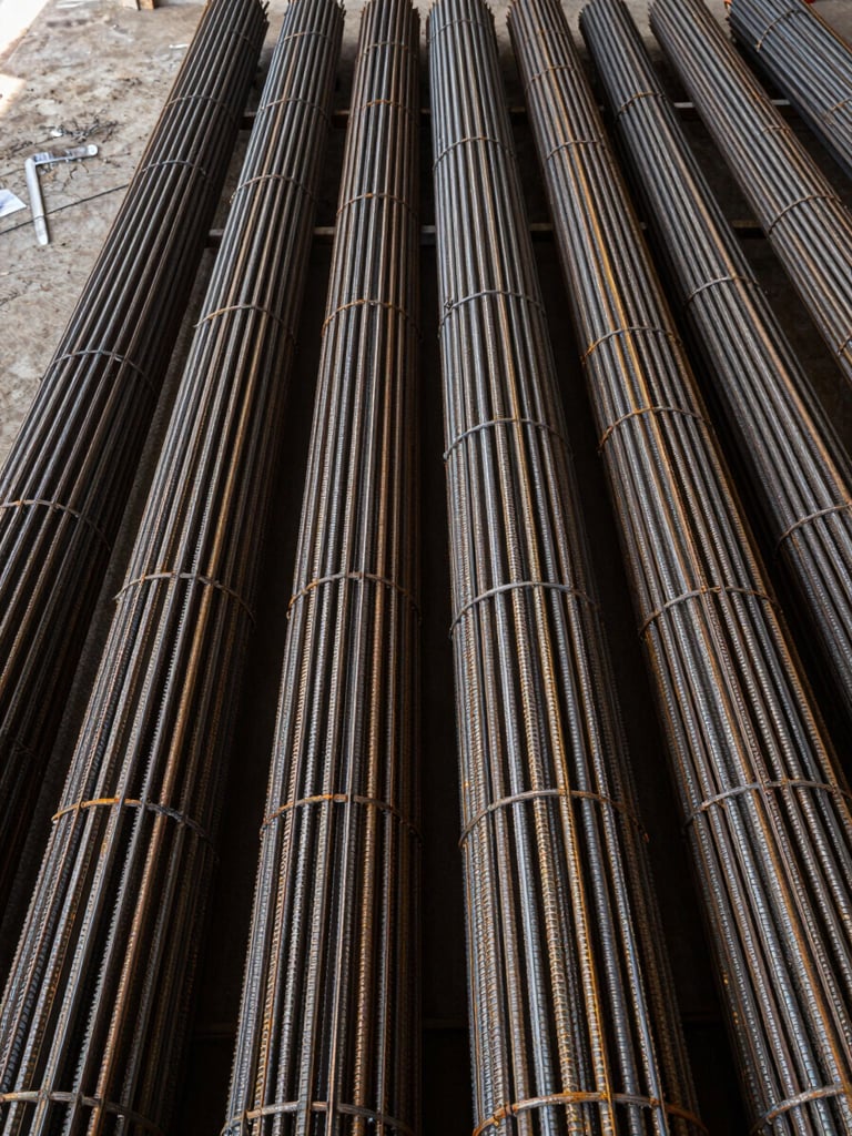 Overhead wide shot looking straight down at tightly packed rebar bundles arranged in a warehouse supply yard, steel surfaces showing mill scale and galvanized sheen, diffused loading-dock daylight from the left edge, no human presence, framing emphasizes repetition and stock depth