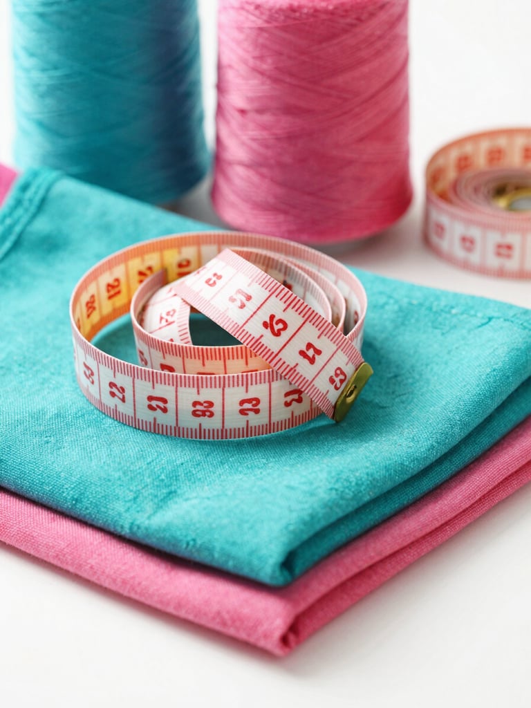 A colorful tailor measuring tape resting on soft folded cotton fabrics, vibrant teal and pink threads in background, bright daylight