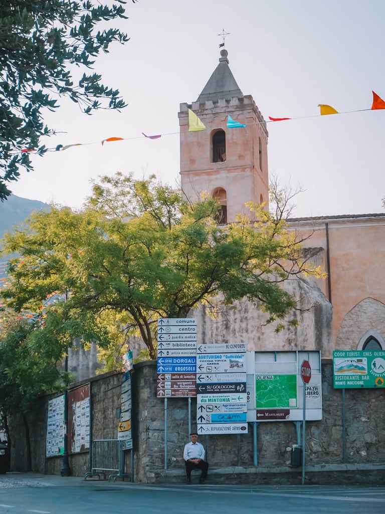 Elderly man in Oliena with street signs and Santa Maria Assunta church tower.