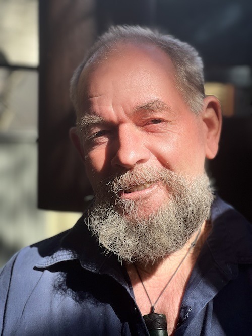 A close-up portrait of a senior man with a grey beard smiling in natural sunlight.