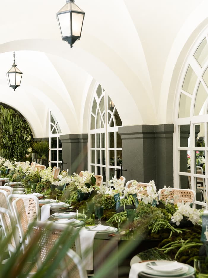 Elegant outdoor dining table setting with green moss runners and white floral centerpieces under arched ceilings.