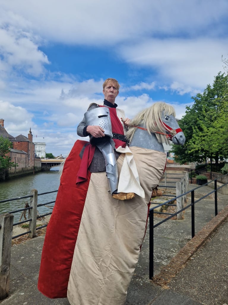 A man in medieval knight armour and red tunic riding a hobby horse stilt walking.