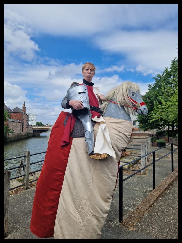 A man in a medieval knight costume riding a pantomime horse by a river in the city.