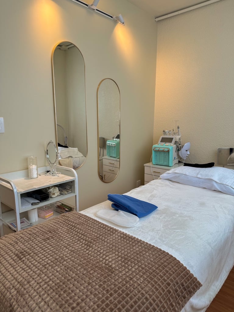 Professional facial treatment room featuring a medical spa bed, oval mirrors, and hydra-facial machine.