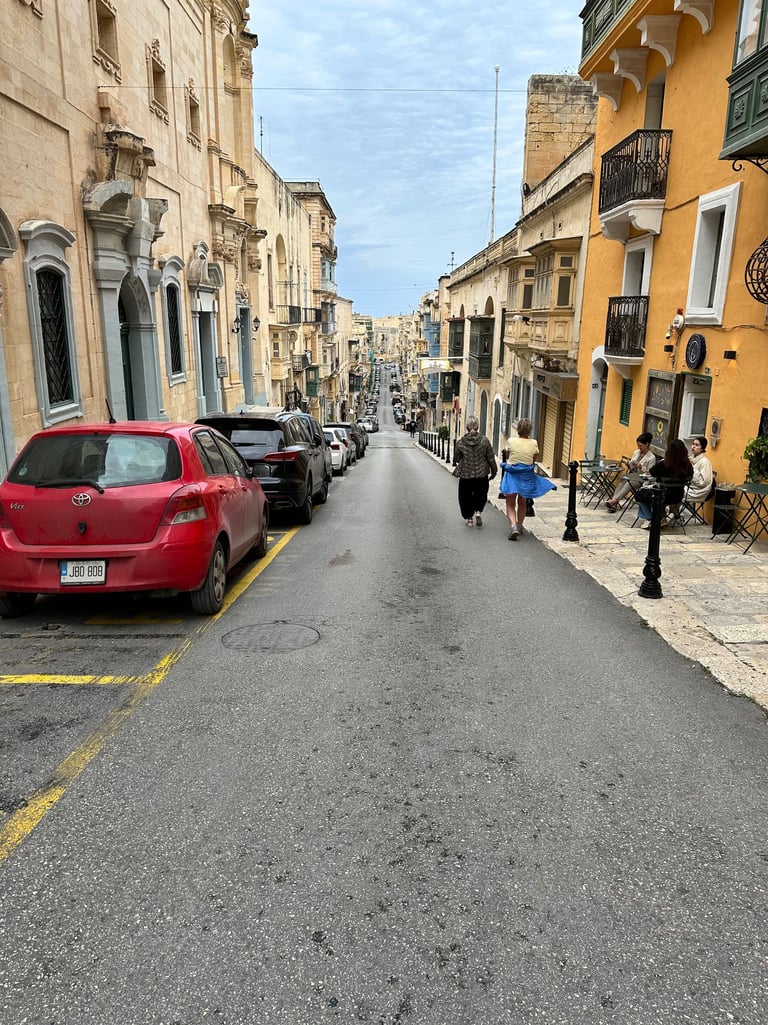 A high-angle view of a narrow, sloping street in Malta, lined with historic buildings and a church o