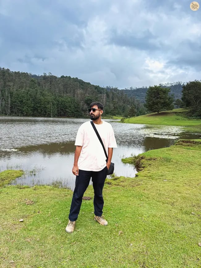 Traveler at the serene shore of Poondi Lake in Kodaikanal, surrounded by calm waters and rolling green hills.