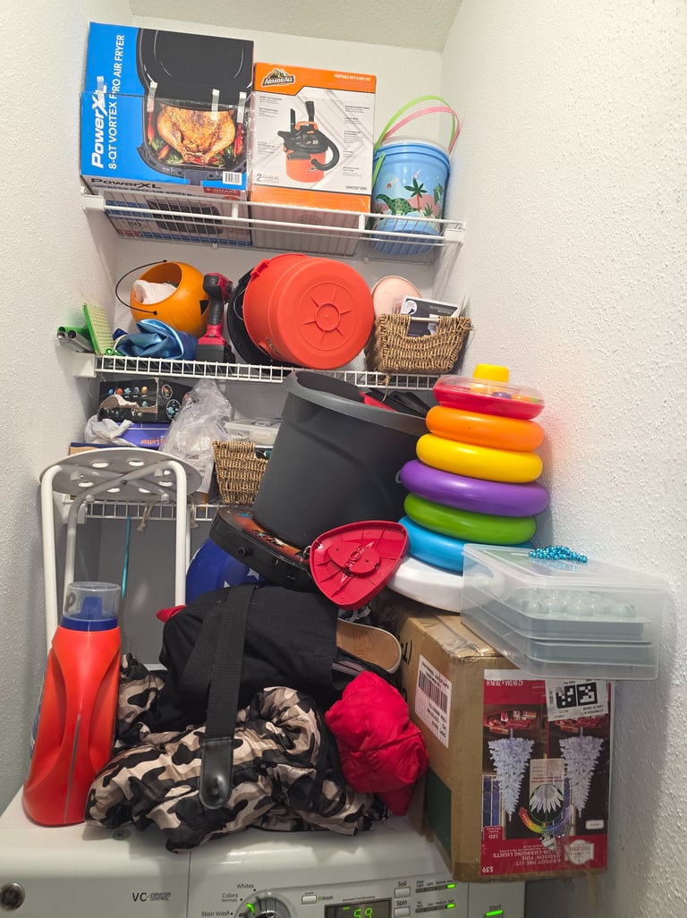 Cluttered laundry closet with shelves holding boxes, toys, and tools above a messy washing machine.