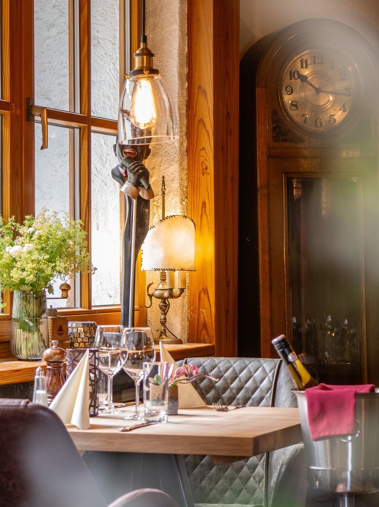 Eleganter Restauranttisch mit Weingläsern am Fenster, antiker Standuhr und warmer Lichtstimmung.