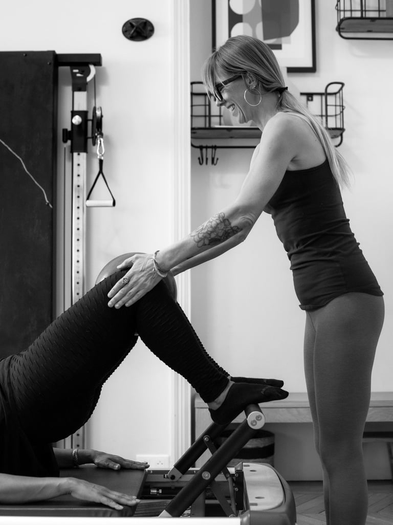 A smiling fitness instructor assists a client with a leg bridge exercise on a Pilates reformer machine.