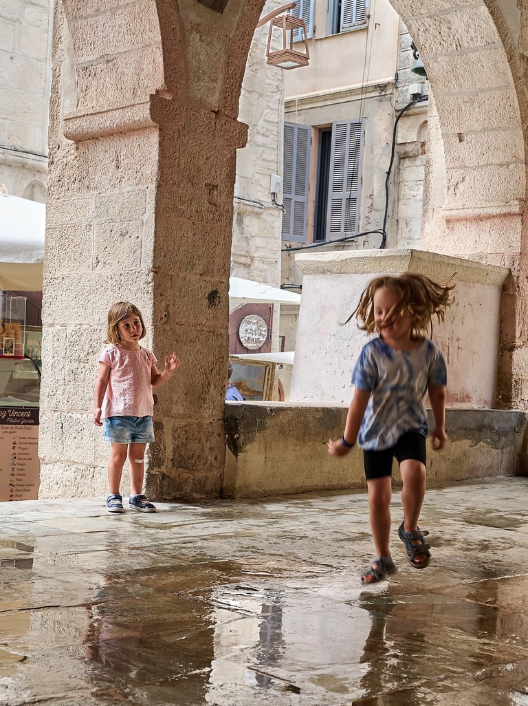 Two young children playing under a historic stone archway on a wet European street.