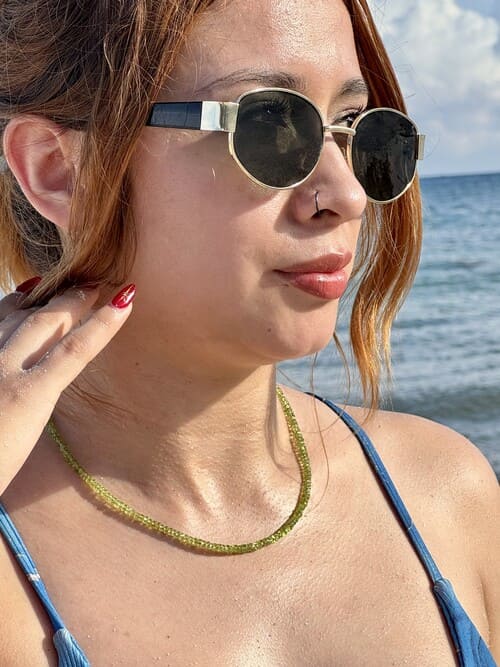 Mujer usando un delicado collar de cuentas de peridoto y gafas de sol redondas en la playa.