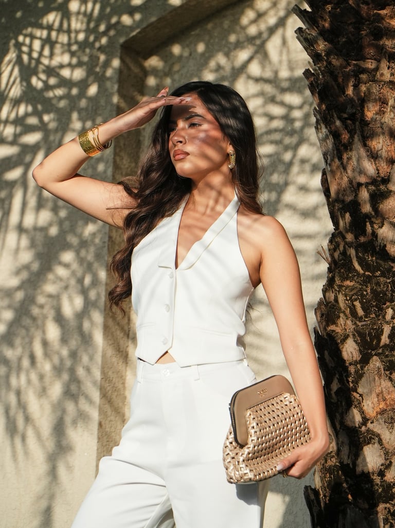 A stylish woman in a white halter vest and trousers poses with a gold woven clutch bag.