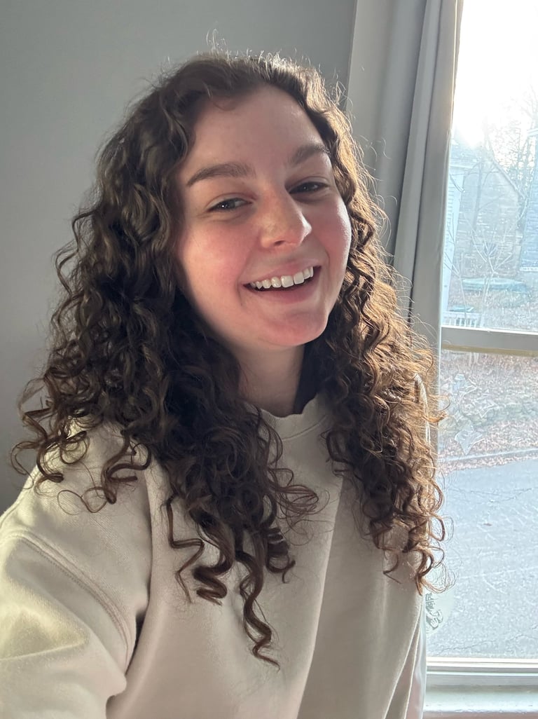Smiling woman with long brown curly hair wearing a beige sweatshirt by a bright window.