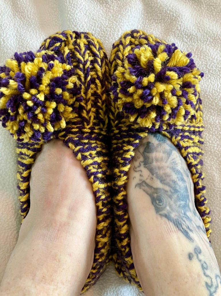 Close up of purple and yellow hand knit house shoes with pompoms on models feet