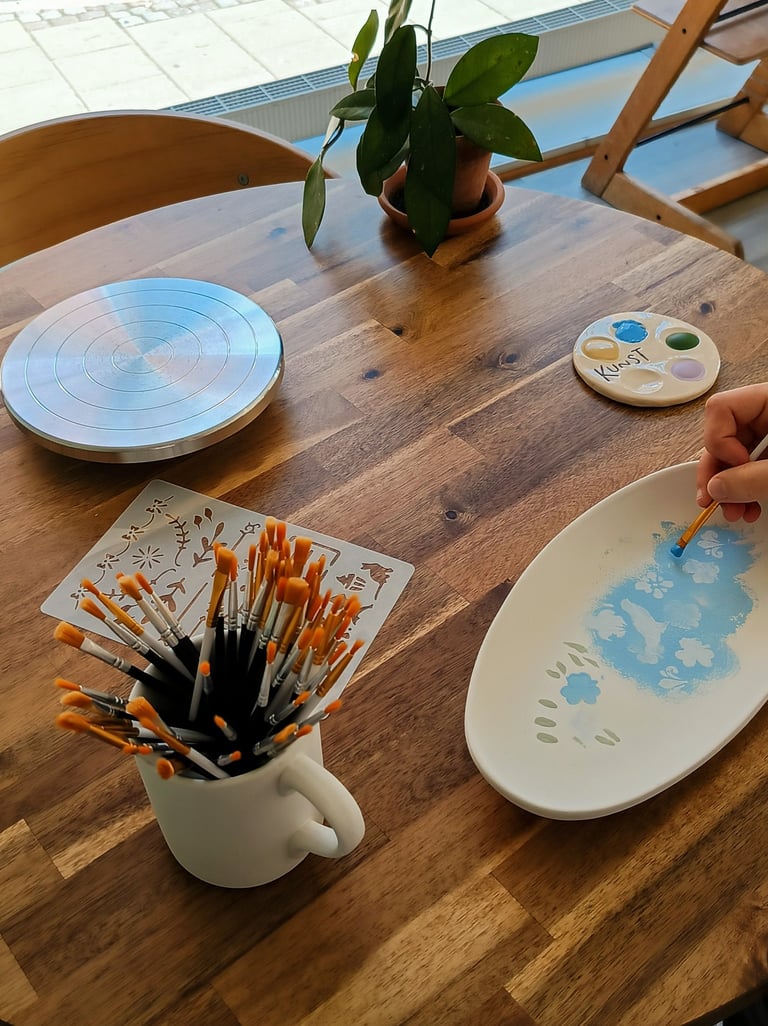 An artist hand paints blue flowers on a ceramic plate using a fine brush at a pottery studio table.