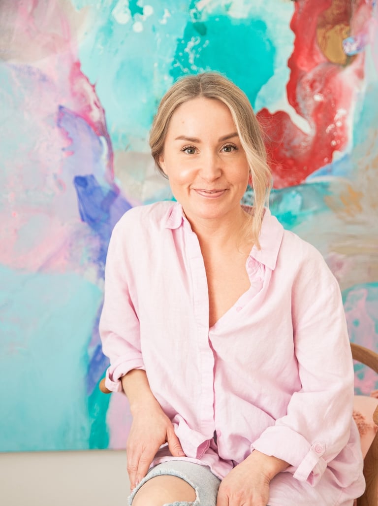 Saija Uussaari in a pink shirt and jeans sitting on a chair in front of a colorful painting