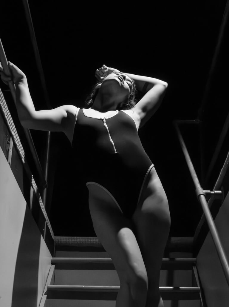 Black and white portrait of a woman in a one-piece swimsuit posing on stairs in dramatic lighting.