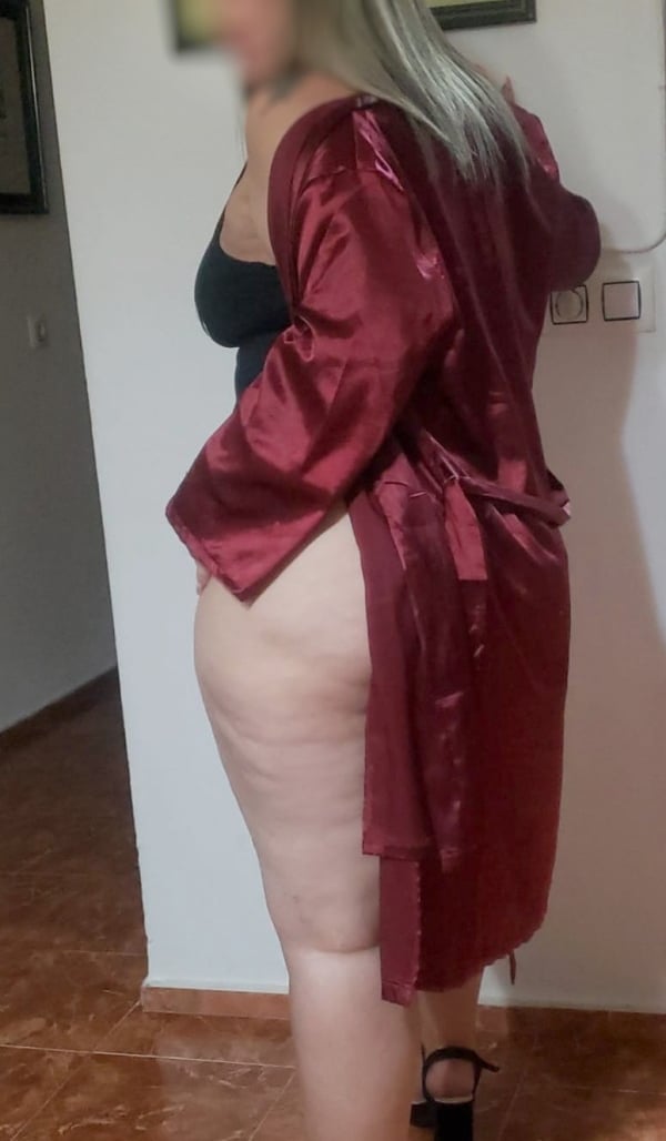 Escort madura colombiana en bata roja de satén, de rodillas sobre el sofá, mostrando sus piernas provocativas.