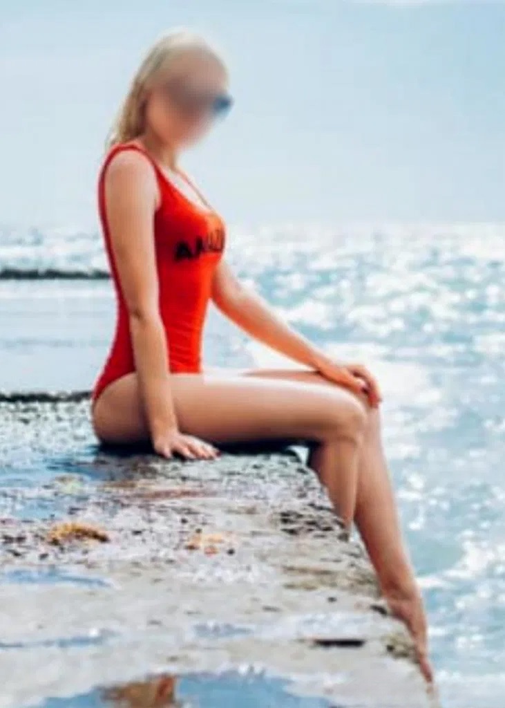 Ana sentada junto al mar en bañador rojo, mostrando su silueta perfecta y su piel dorada.