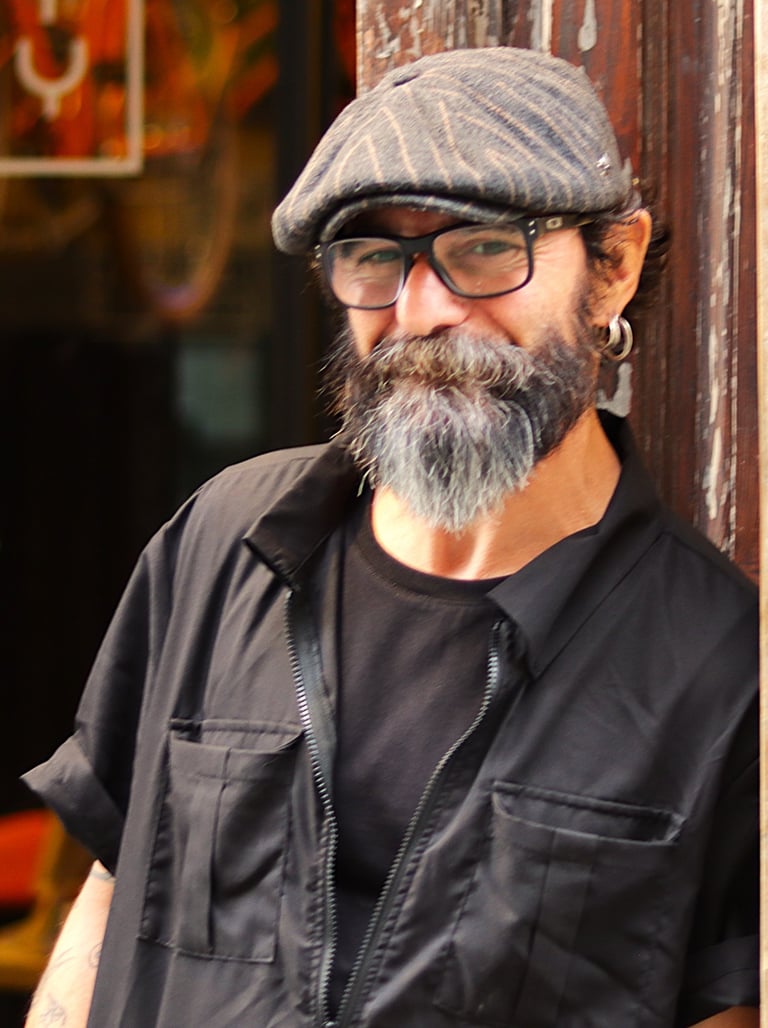 Smiling gray-bearded man in glasses and flat cap stands outdoors by orange URBAN sign, blue sky behind. Black shirt and jacke