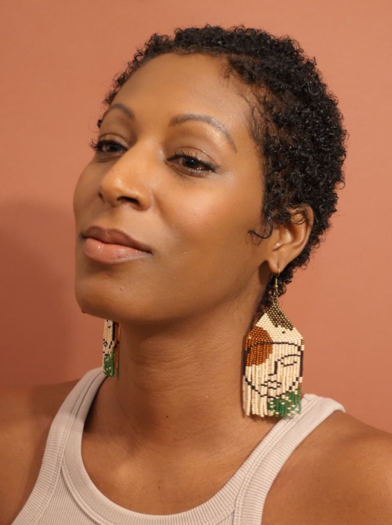 A woman with a white tank top wearing a pair of  earrings beaded by hand with a face pattern