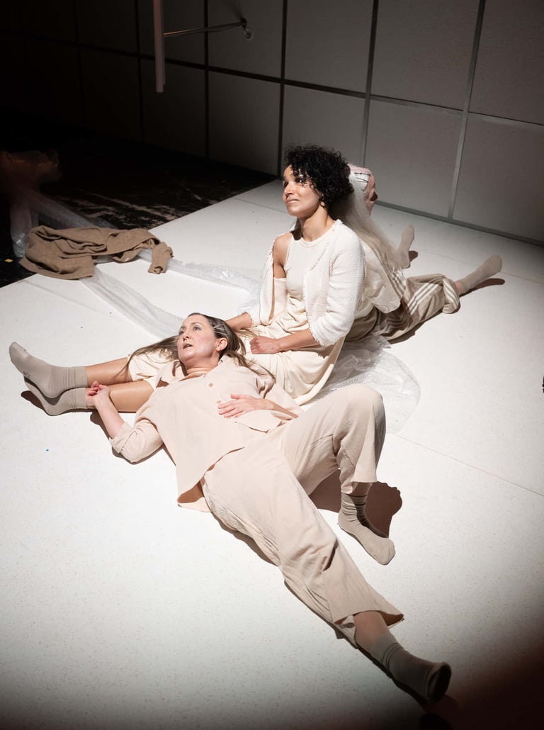 Eine Theaterszene. Drei hell gekleidete Frauen sitzen einträchtig auf dem Boden.