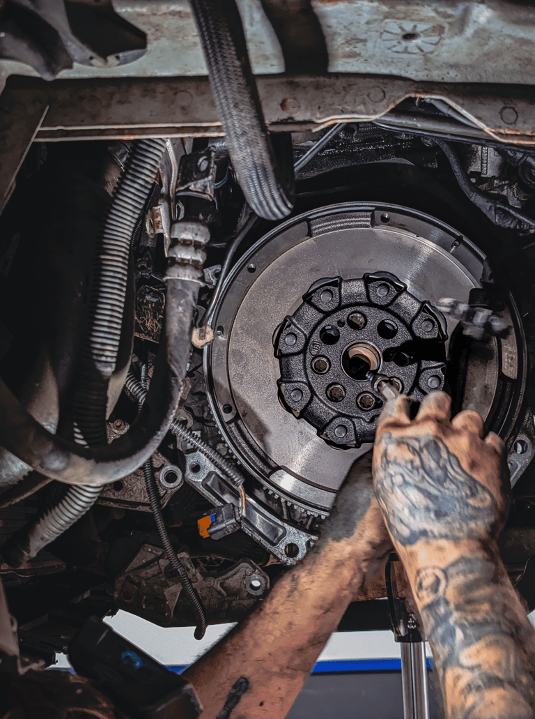 A mechanic with tattooed arms repairs a car engine flywheel and clutch assembly in an auto shop.