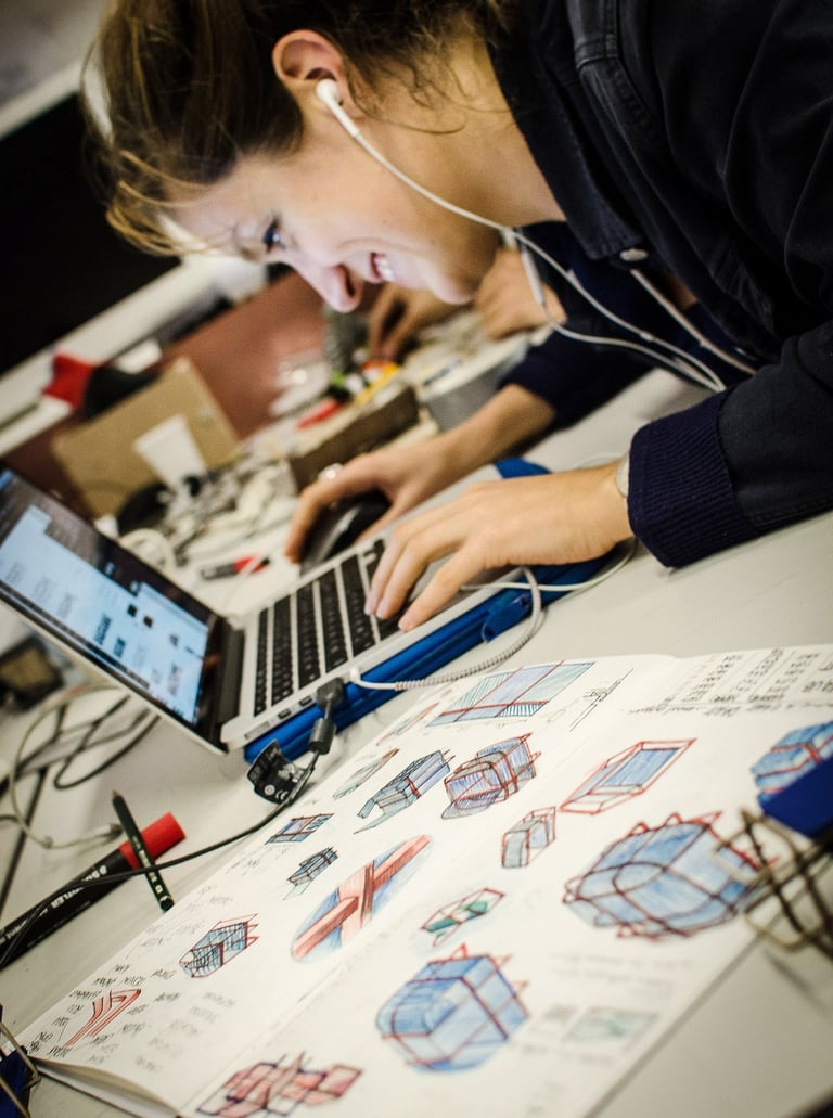A design student sketching industrial concepts next to a laptop in a creative workspace.