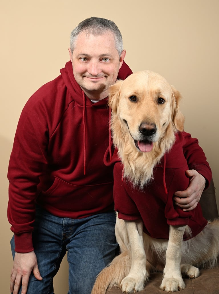 A smiling man and his Golden Retriever wearing matching maroon hoodies in a studio portrait.