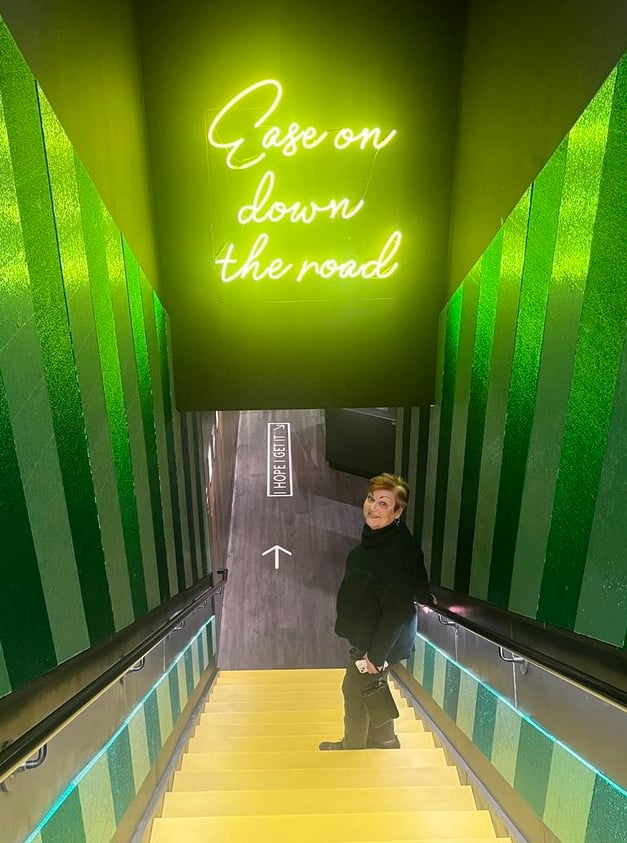 Photo of Debbie going down yellow stairs with green striped walls, standing before a neon sign: "Ease on down the road."