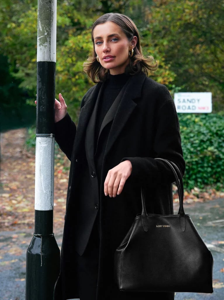 A stylish woman in a long black wool coat carries a luxury leather tote bag on a street.