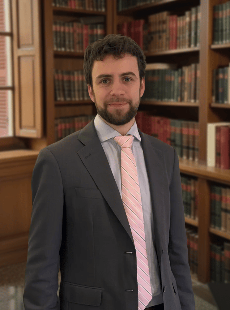 Professional portrait of a lawyer in a charcoal suit and pink tie standing in a law library.