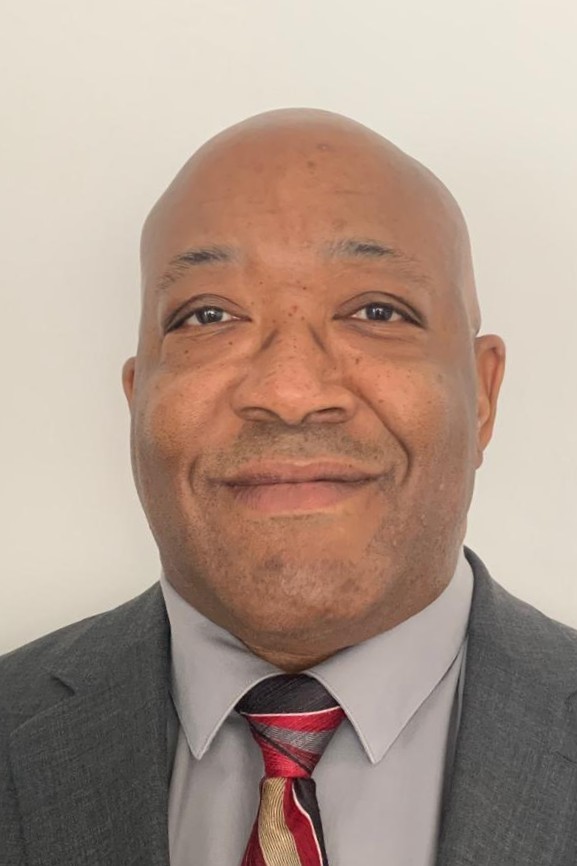 Professional headshot of a smiling bald Black man wearing a grey suit jacket and a striped necktie.