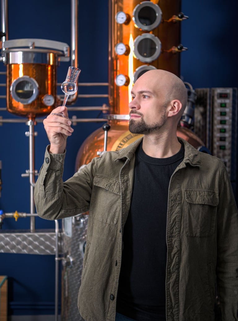 mastro distillatore che osserva del liquido in un bicchiere da degustazione davanti a un alambicco