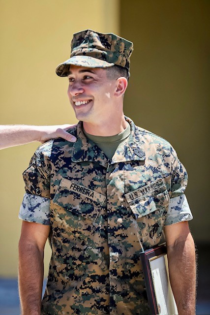 A smiling US Marine in MARPAT camouflage uniform holding a framed award certificate at a ceremony.