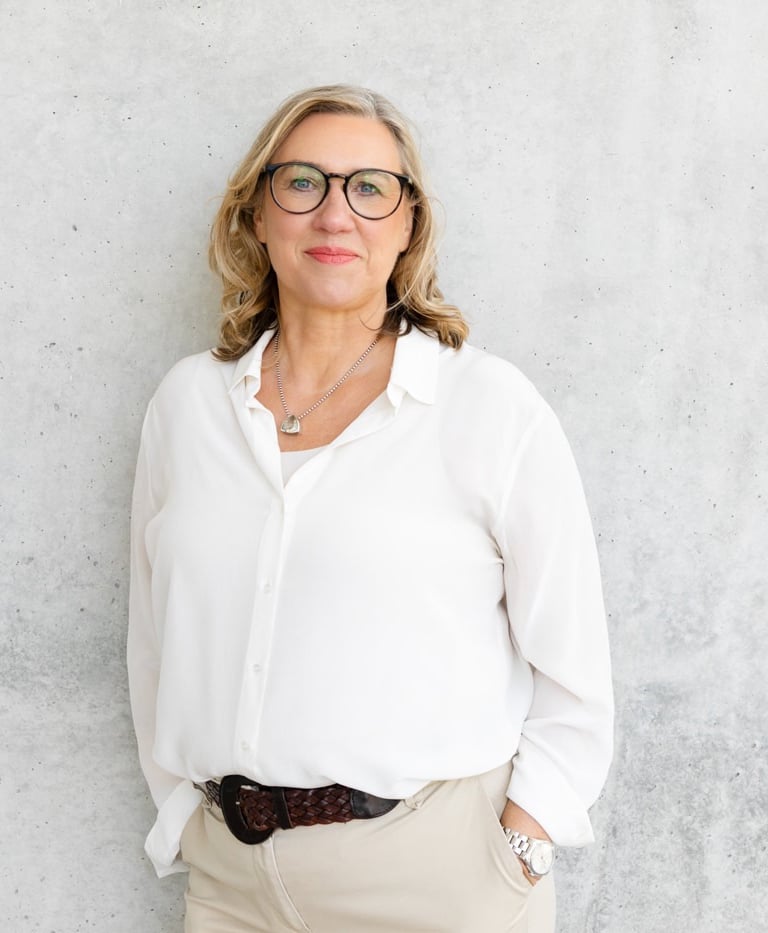 Gundula steht in weißer Bluse und Brille selbstbewusst und locker vor einer weißen Betonwand.