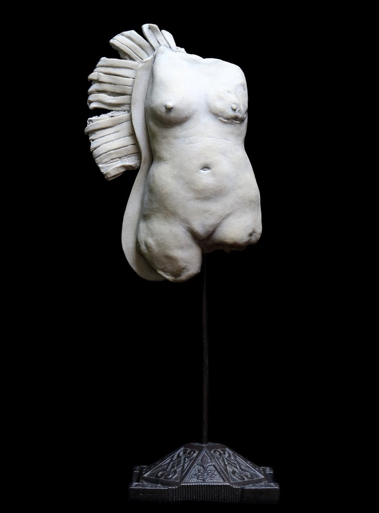 Small ceramic fragment of a nude female figure with a disfigured breast and bedraggled, wing-like fringe.