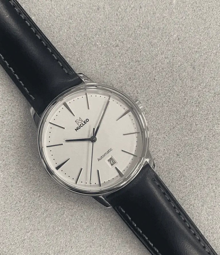 orologio dress nucleo con quadrante bianco indici e lancette argento data a ore 6 cinturino pelle nera posto su piano grigio