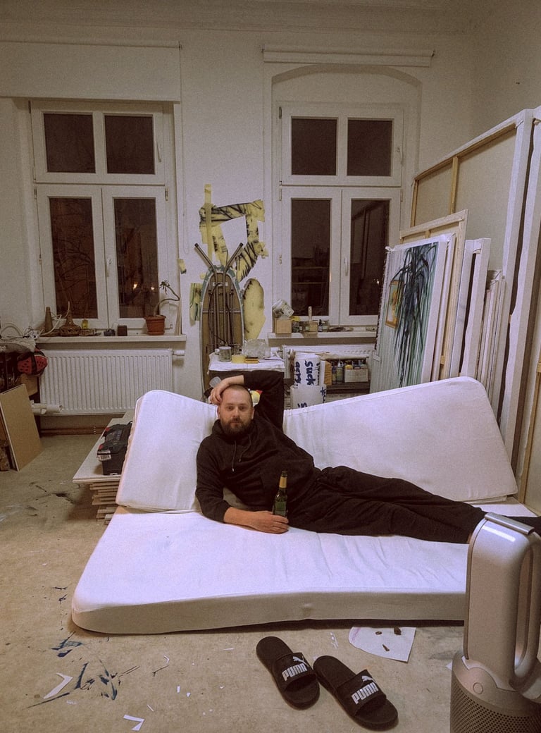 Artist relaxing on a floor mattress in his messy creative studio with paintings