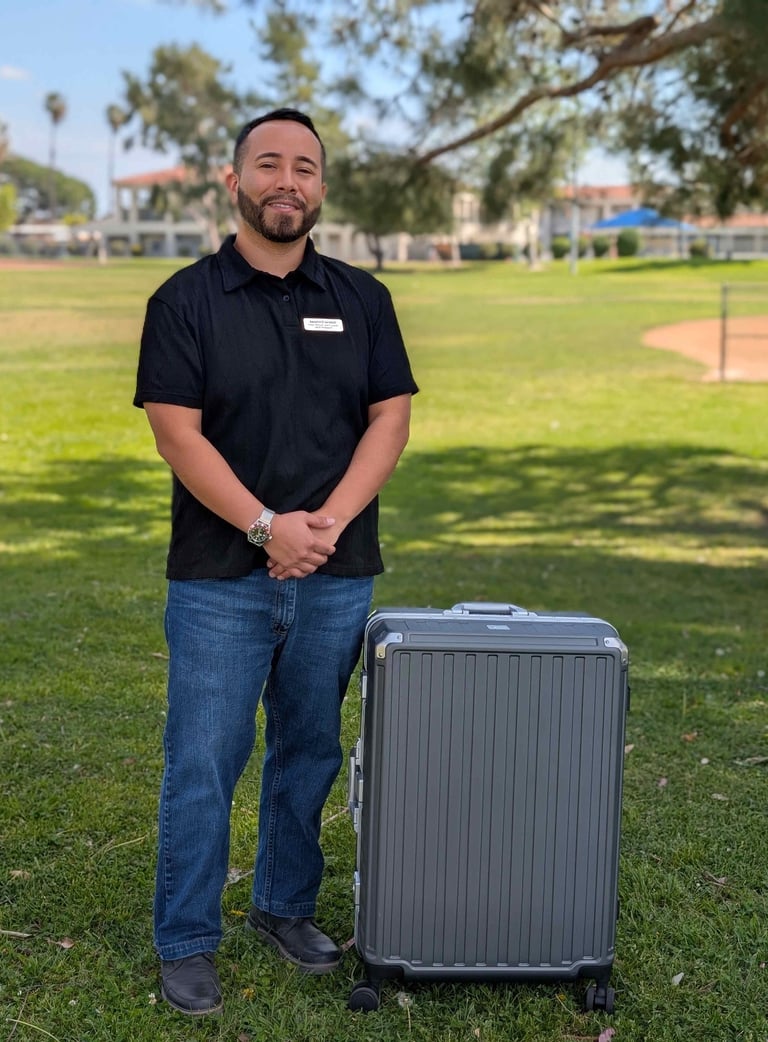 A smiling man standing in a park next to a large silver hardshell suitcase on wheels.