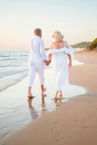 Pareja de adultos mayores vestida de blanco caminando de la mano por una playa de arena al atardecer
