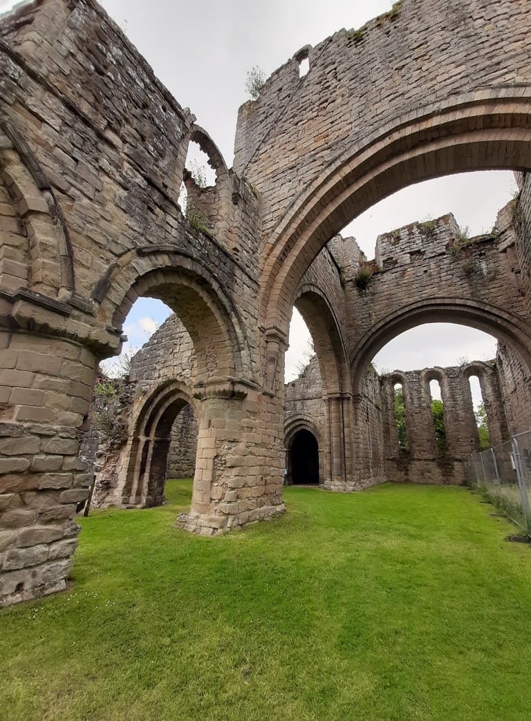 Ancient stone ruins of a medieval abbey with gothic arches and grassy interior under a cloudy sky.
