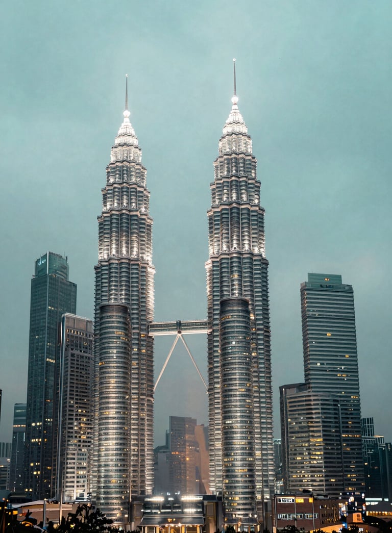 A wide-angle professional photograph of the Kuala Lumpur skyline featuring the Petronas Twin Towers at dusk. The sky is a soft mist and pale aqua. The city lights start to twinkle, reflecting muted slate blue tones. High-end, clean architectural photography.