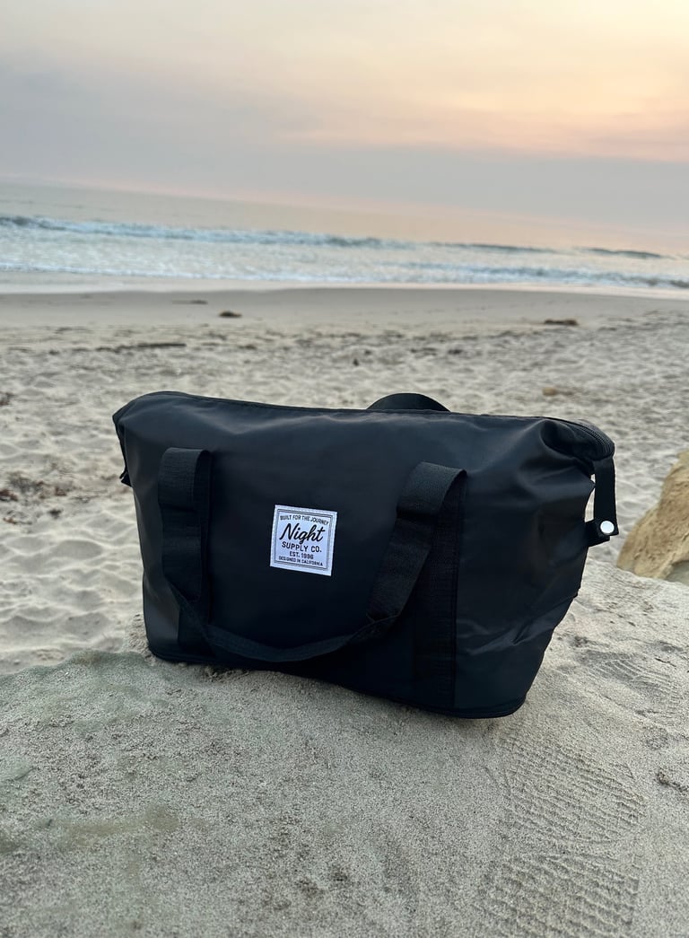 Black travel bag from Night Supply Co. placed on a sandy beach at sunset, with ocean waves and mount