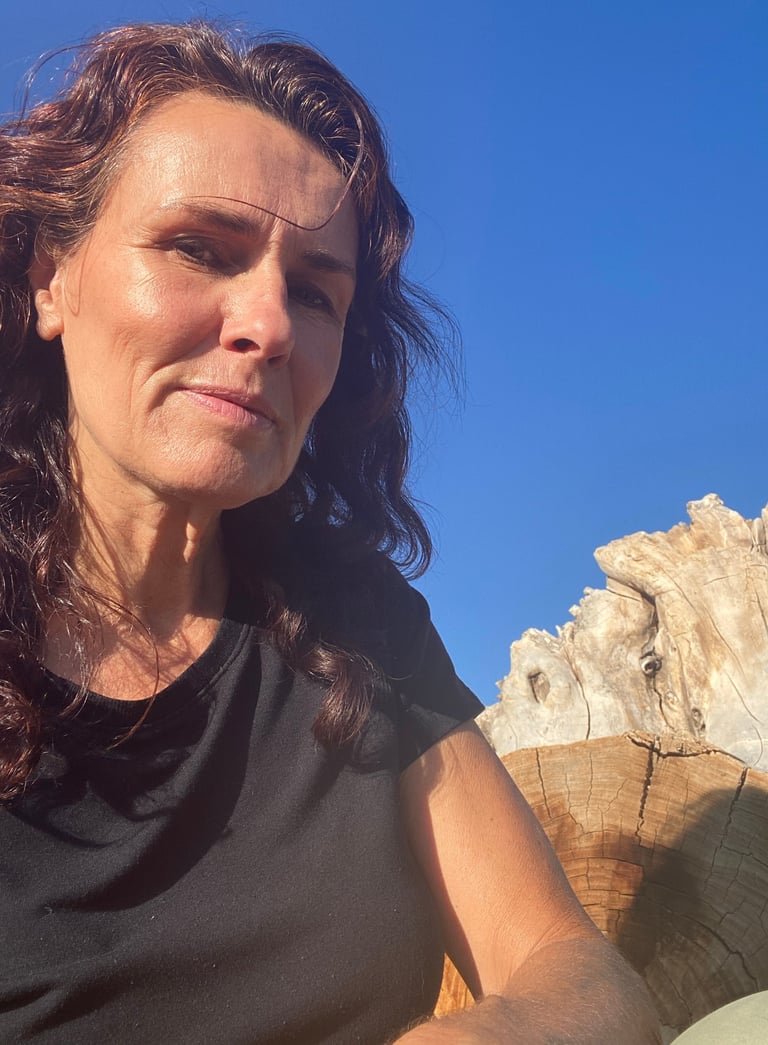 A woman with wavy brown hair posing outdoors near weathered driftwood against a clear blue sky.