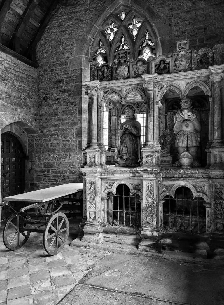 Black and white interior of a stone church featuring ornate gothic tombs, statues, and a wooden cart.