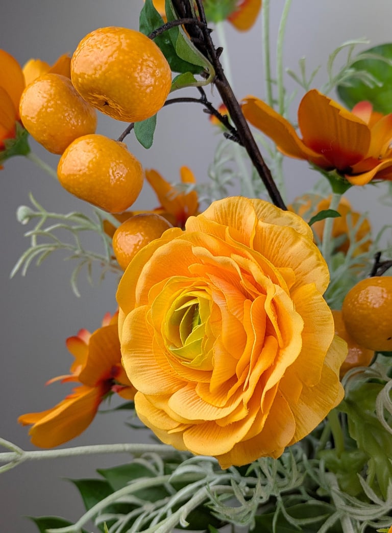 Artificial orange ranunculus flower arrangement with decorative faux citrus fruits and green leaves.