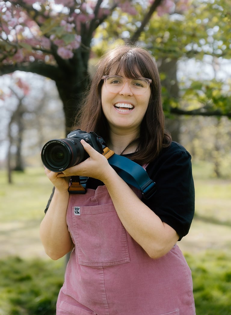 Laura Franklin holds her camera up as she smiles to the camera. It is a London Blossoms photo shoot 