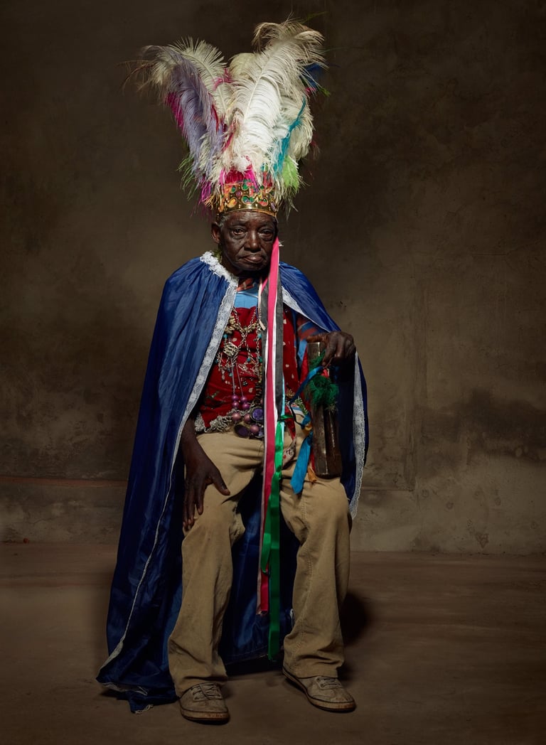 An old black man in a ritualistic gound with a feathered headdress, cape in a rugged cement backgrou